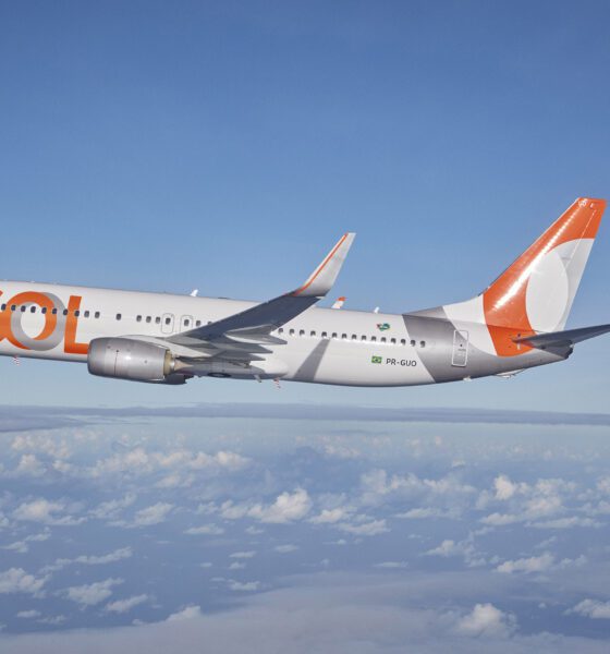 Avião da Gol voando no céu azul, com nuvens abaixo.