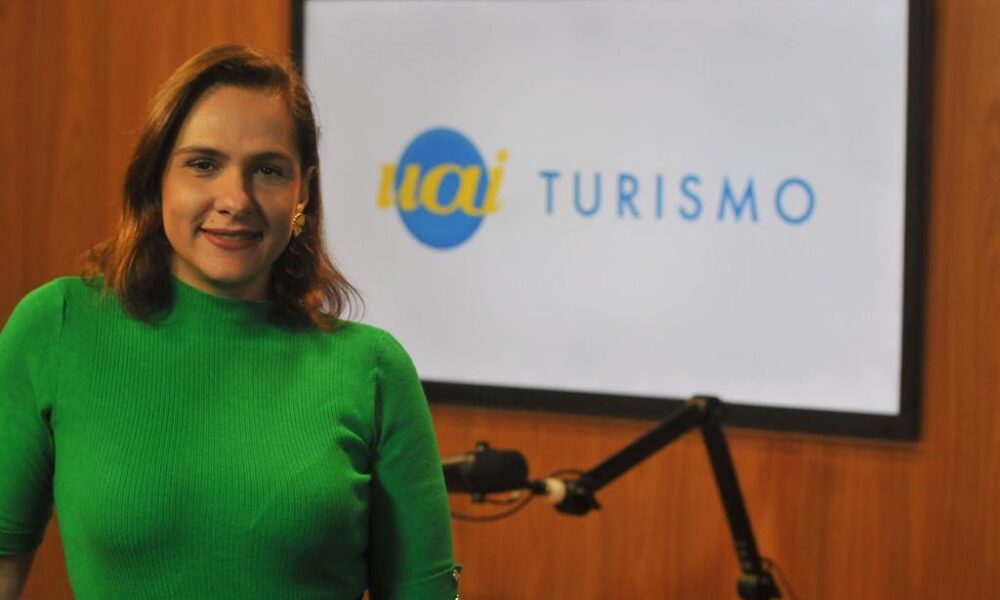 Mulher adulta morena, de blusa verde e ao fundo, uma televisão exibe a marca Uai Turismo