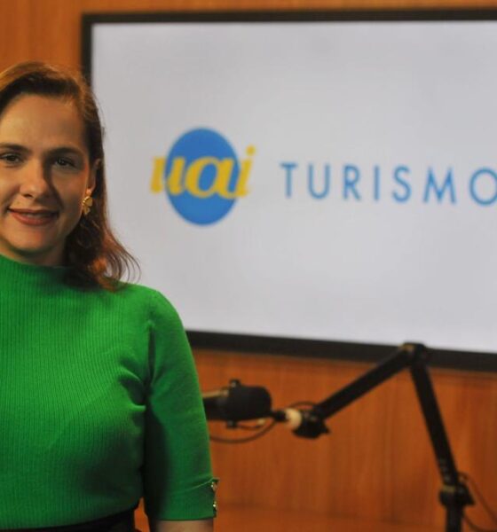 Mulher adulta morena, de blusa verde e ao fundo, uma televisão exibe a marca Uai Turismo