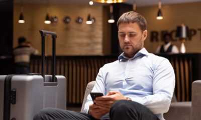 Homem branco de roupa social sentado em recepção de hotel, mexendo no celular. Uma mala cinza está ao lado dele.