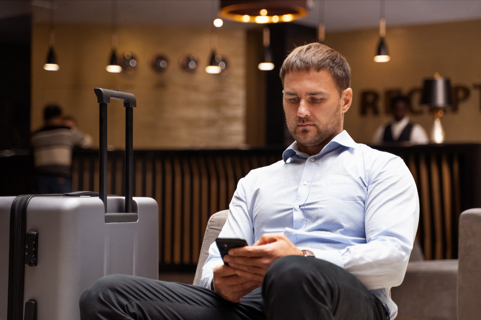 Homem branco de roupa social sentado em recepção de hotel, mexendo no celular. Uma mala cinza está ao lado dele.