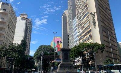 Foto de Belo Horizonte, centralizando o Obelisco da Praça 7 de setembro. Atrás aparece a pintura "Deus é mãe" do artista Robinho Santana, que representa uma mãe negra com dois filhos, um no colo e outro em pé ao seu lado.