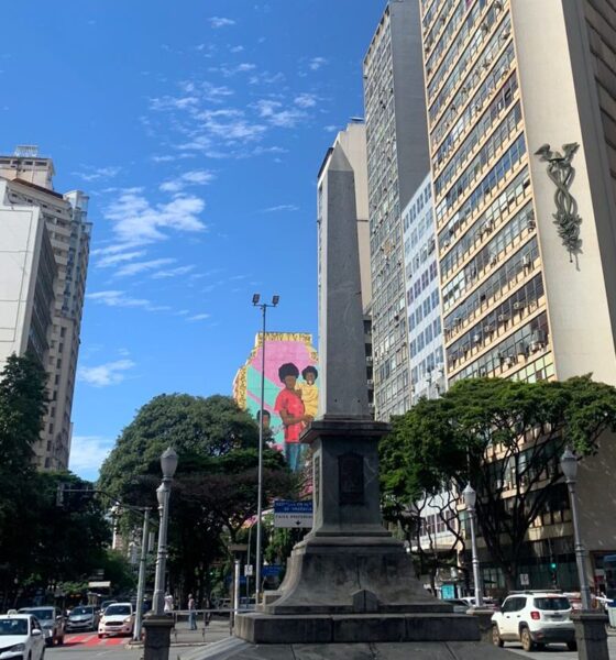 Foto de Belo Horizonte, centralizando o Obelisco da Praça 7 de setembro. Atrás aparece a pintura "Deus é mãe" do artista Robinho Santana, que representa uma mãe negra com dois filhos, um no colo e outro em pé ao seu lado.