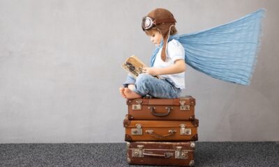 Criança vestida de aviador com um livro nas mãos, em cima de 3 malas empilhadas