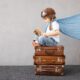 Criança vestida de aviador com um livro nas mãos, em cima de 3 malas empilhadas