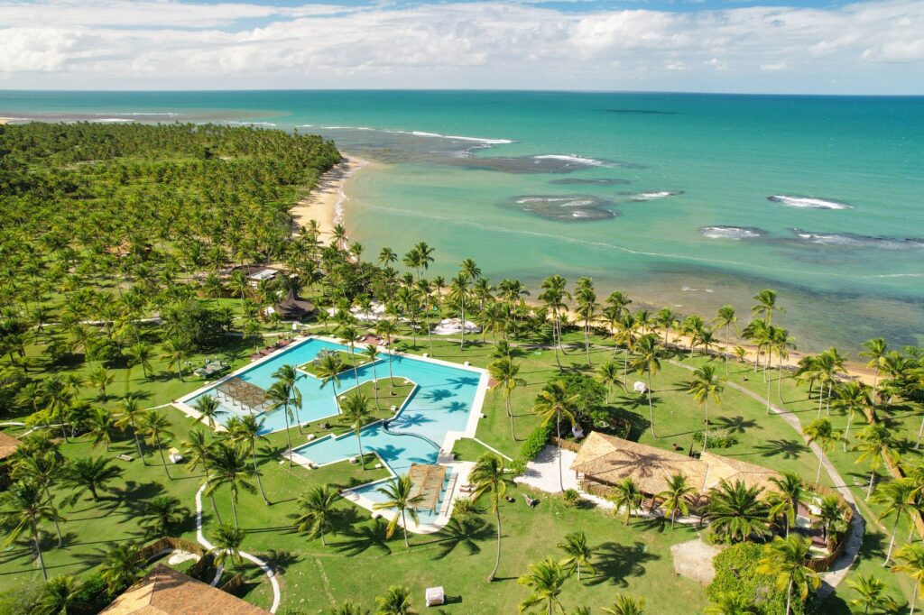 Vista aérea do hotel de frente para o mar azul, com área verde com muitos coqueiros